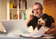 Older man talking on phone looking at laptop and sitting with dog on his lap